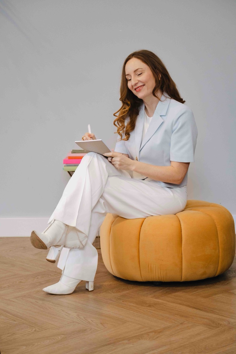 lucinda allen smiling and writing in a notebook, sitting on a mustard velvet pouf